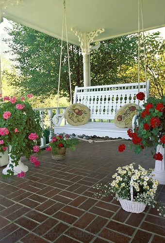 Brick tile front porch with hand made victorian wood painted swing with two hooked pillows and many potted plants.