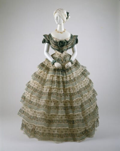 An 1855 evening ensemble with an elaborate 7-layer flounced skirt decorated with flowers and stripes. The introduction of the cage crinoline in the 1850s allowed skirts to reach enormous sizes. It als…