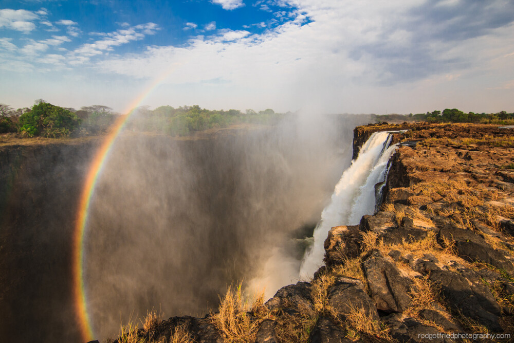 A rainbow forms as the morning sun hits the spray coming off Victoria Falls. This photo was taken from the Zambian side of the falls, on the way to visit Devil's Pool. In high season the entire rocky area is covered by flowing water.