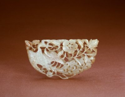 玉孔雀衔花饰，宋，长7.6cm，宽3.8cm。清宫旧藏。 花饰玉色青白，有赭色斑，为半圆形玉片，其上透雕孔雀衔花图案。图案主体为孔雀，孔雀回首，拖尾，展翅，口衔花枝，枝上有花两朵，品种不同。 花鸟类玉器在…