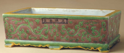 明正德   素三彩缠枝莲纹长方水仙盆，明正德，高7.2cm，口径23.8cm×15.2cm，足径23.1cm×14.2cm。 盆呈长方体，四面略斜收，下承以六足。盆内及外底均施白釉。外壁素三彩装饰，以黄、绿、紫设色，腹部以紫色为地…