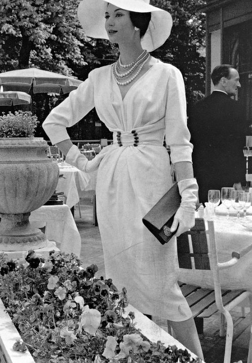 1957 Simone d'Aillencourt in white crépe dress with softly draped bodice cinched by buckled waistband by Nina Ricci,