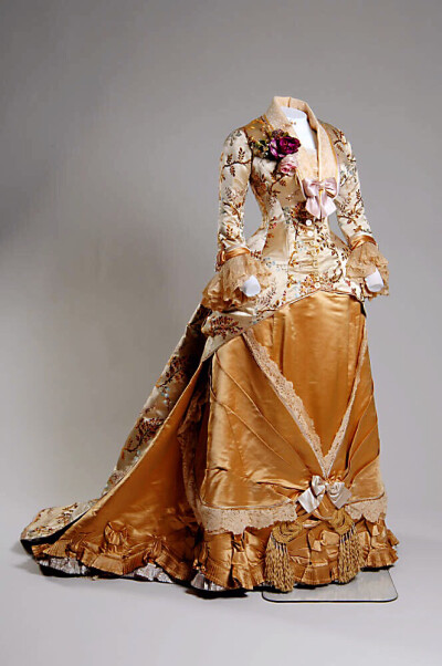 1878 Dinner dress 白色锦缎底子遍布藤蔓与花朵，袖口和领子饰以蕾丝和丝缎蝴蝶结。胸前别着粉紫色小花束。蕾丝饰带将金色缎子裙切割成不同的三角形区域，汇聚在裙子底部缎带蝴蝶结处，再垂下流苏装饰。