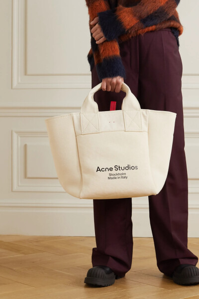 Acne Studios 这款手提包容量颇大，适合在长途通勤或是短期旅行时使用。单品采用纯棉帆布在意大利制成，印有黑色的品牌名称，开放式包口配有红色皮革袢带。不妨使用带拉链的口袋稳妥放置银行卡或钥匙，以免它们落入…