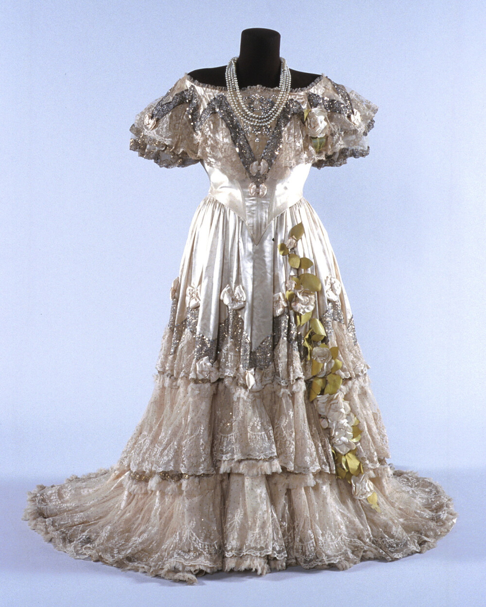 澳大利亚歌剧演唱家Nellie Melba在1900年主演《茶花女》时穿的戏服，由当时著名的时装师Jacques Doucet设计。象牙色缎子裙，装饰着层叠的蕾丝花边、大量银色亮片与水钻组成的闪光饰带、缎子蝴蝶结，还有布制白色山茶花，让整个人都包裹在一片柔光中。款式结合了故事所在的1840年代的服装与演出当时的流行风格，这也是过去的舞台服装常用的设计手法。