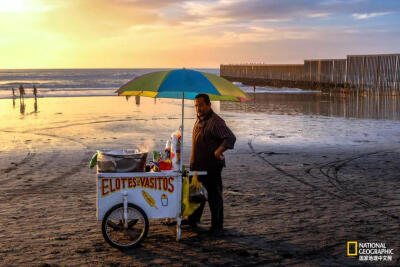 海滩小贩
在墨西哥和美国边境的Playa de Tijuana，一名小贩向海滩游客兜售“elotes”(一种带酸橙汁、奶油、奶酪和香料的玉米棒)。摄影：SETH BERRY