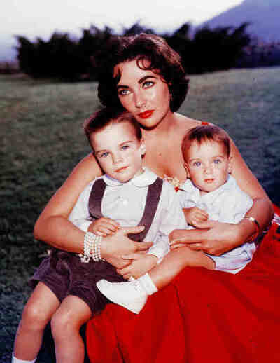 Elizabeth Taylor sits in a red evening dress  with her sons Michael and Christopher Wilding on her lap, mid 1950’s.  ​​​