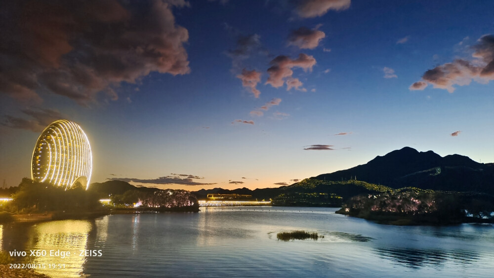 在北京怀柔区的雁栖湖边，吹着夜风，赏着夜景，波光粼粼，落日余晖就像颜色滤镜一般过渡柔和，云朵非常配合，飘在了合适的位置。