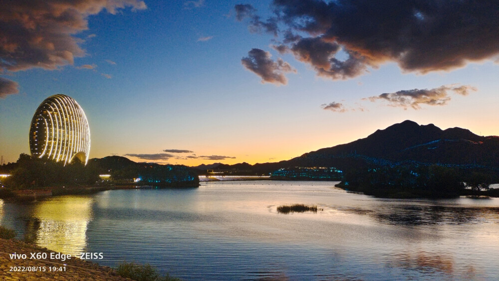 在北京怀柔区的雁栖湖边，吹着夜风，赏着夜景，波光粼粼，落日余晖就像颜色滤镜一般过渡柔和，云朵非常配合，飘在了合适的位置。