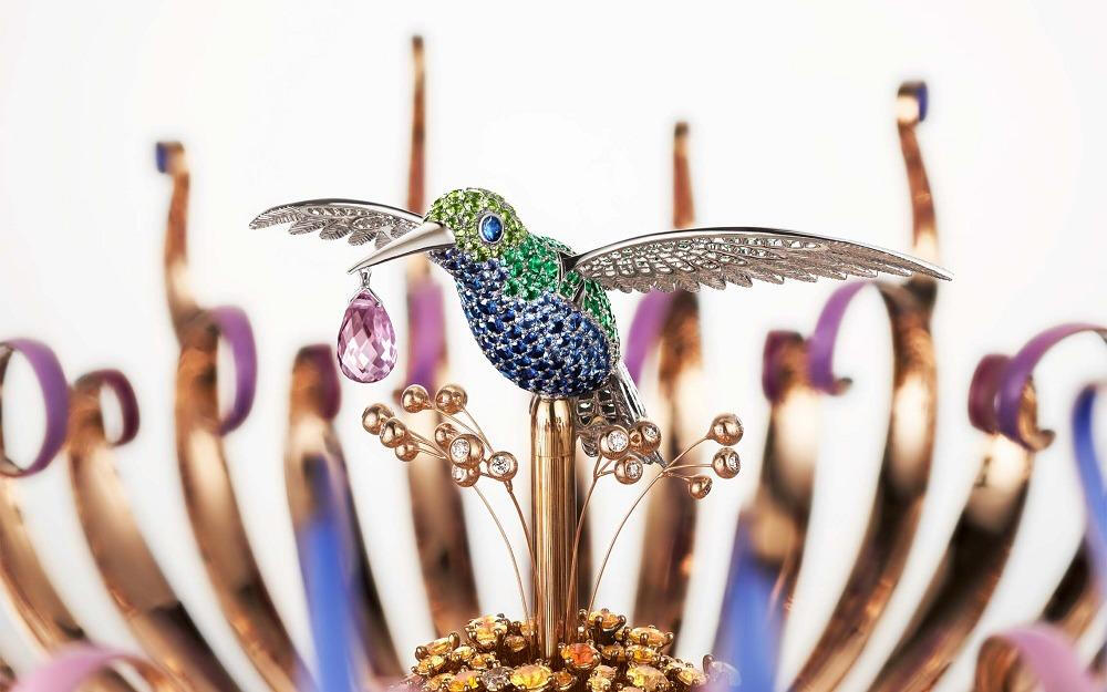 梵克雅宝“Van Cleef & Arpels”在伦敦巨匠臻藏艺术博览会（Masterpiece London）上展出 Rêveries de Berylline“绿蜂鸟之梦”自动机械装置，“Rêveries de Berylline”是一座独一款自动机械报时装置，延续梵克雅宝（Van Cleef & Arpels） 的自动机械人偶传统，构筑出蜂鸟在花间盘旋的梦幻场景——蜂鸟轻快扑打翅膀，口中衔着一颗露珠，紫粉色花瓣层层绽放，充满浪漫诗意。
整座装置直径为21.5cm，高度为27cm，底座由红斑岩雕刻而成，外圈环绕着金质刻度环，以一片镶钻金叶来指示当前时间；底座上方可以看到碗状青金石托起的梦幻场景，轻轻转动发条启动报时装置，可以看到合拢的花瓣缓缓展开，蜂鸟随着清脆的报时音展翅飞舞，稍稍盘旋后悠悠飞回花冠中央。
设计师以白金塑造出蜂鸟玲珑的身形，斑斓羽翼由蓝宝石、祖母绿、沙弗莱石交替铺排，鸟喙下方还垂落着一颗水滴形淡紫色蓝宝石，随蜂鸟的动作晶莹颤动；卷曲的花瓣饰以紫粉色漆面，展现柔美而细腻的色彩渐变；花蕊则由橙色石榴石、黄色蓝宝石、钻石锦簇而成，与一侧垂落的金质叶片相映成辉。