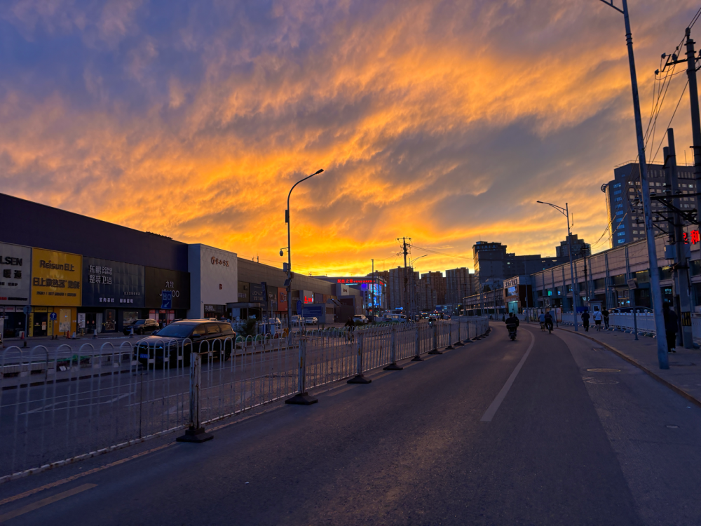 猫哥记录：日落下的城市，仿佛一幅生动的油画！
天空中，橙黄渐变的云层如同温柔的晚霞，预示着太阳即将拥抱大地。街道两旁，现代建筑矗立，广告牌闪烁着绚丽的光芒，为这宁静的傍晚增添了几分活力。
街道上，车辆已悄然离去，只留下几辆静静停放的汽车。一位行人悠闲地走在人行道上，深色衣服与周围景色形成鲜明对比，仿佛他是这画中的主角。
电线杆和路灯整齐排列，发出柔和的光芒，与天空中的晚霞相互辉映。整个场景充满了宁静与平和，仿佛时间在这一刻都放慢了脚步。
此刻的我，只想静静欣赏这美丽的景色，感受这份难得的宁静与美好。