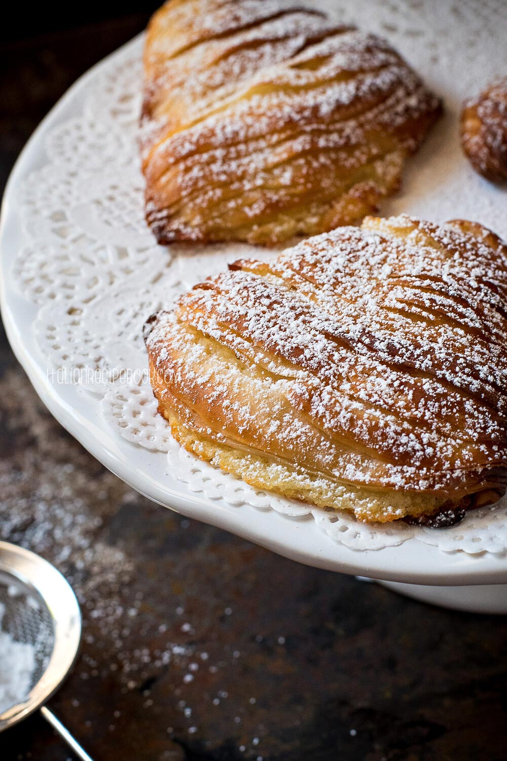 意大利甜品 Sfogliatella 意式千层酥 夹心奶酪千层酥，在意大利语中意为“层层叠叠”“龙虾尾”，指的是蛤蜊贝壳的层状结构，经过烘烤又香又脆。据说最初的千层酥是1700年左右阿马尔菲海岸的修女们发明的，虽然修女们被禁止与外界接触，但配方还是被一位那不勒斯厨师莫名其妙地得到了，这款甜点很快便成为那不勒斯人的最爱。外皮做工非常复杂，需要好几个人在一张很大的工作台上把每层面皮拉到极薄。内馅由乳清干酪、蛋奶冻、杏仁霜、橙皮、水果蜜饯等一系列极具意大利特色的材料混合而成，充满了嫩滑甜蜜。撒上糖霜，趁热品尝外皮尤其酥脆可口