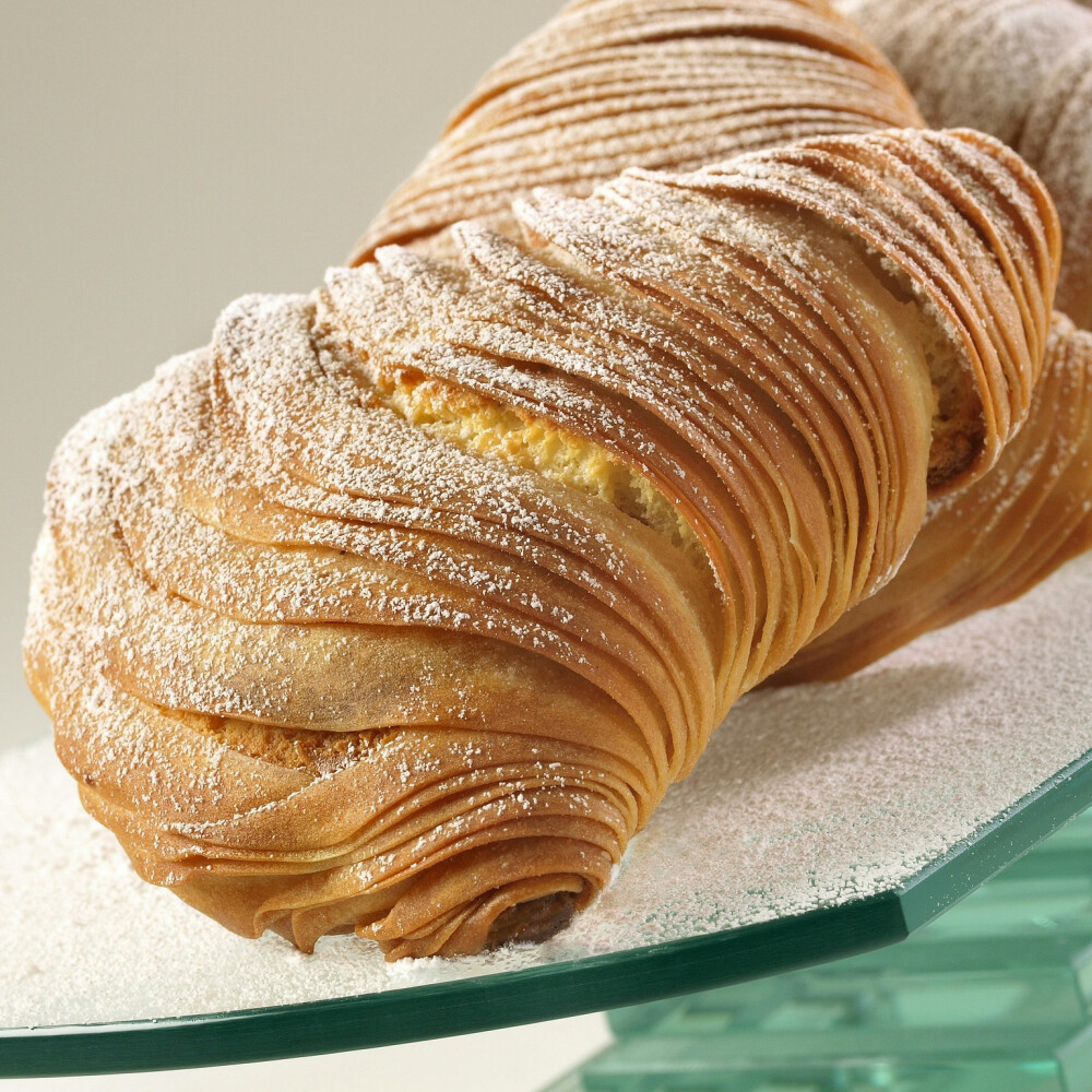 意大利甜品 Sfogliatella 意式千层酥 夹心奶酪千层酥，在意大利语中意为“层层叠叠”“龙虾尾”，指的是蛤蜊贝壳的层状结构，经过烘烤又香又脆。据说最初的千层酥是1700年左右阿马尔菲海岸的修女们发明的，虽然修女们被禁止与外界接触，但配方还是被一位那不勒斯厨师莫名其妙地得到了，这款甜点很快便成为那不勒斯人的最爱。外皮做工非常复杂，需要好几个人在一张很大的工作台上把每层面皮拉到极薄。内馅由乳清干酪、蛋奶冻、杏仁霜、橙皮、水果蜜饯等一系列极具意大利特色的材料混合而成，充满了嫩滑甜蜜。撒上糖霜，趁热品尝外皮尤其酥脆可口