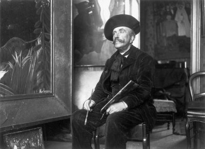1907 年，亨利·卢梭坐在巴黎佩雷尔街 2 号之二的工作室里，旁边是他的画作《被老虎袭击的童子军》。照片由多尔纳克（保罗·弗朗索瓦·阿诺德·卡东；法国，1859–1941 年）拍摄。