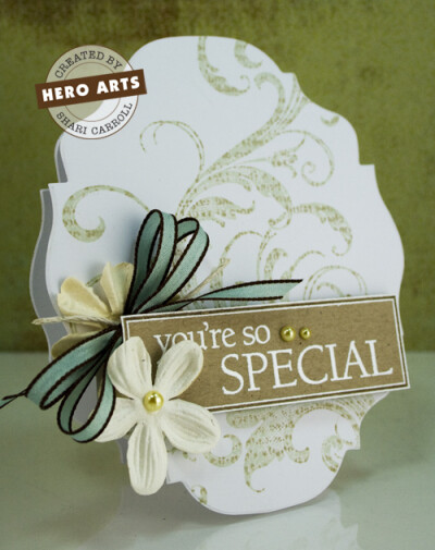 Kissing with Two Inked Stamps Die cut a piece of Snow Layering Paper to create a decorative tag shaped card. Ink the Fabulous Flourish stamp with Soft Leaf Shadow Ink. Ink the Friends Definition stamp…