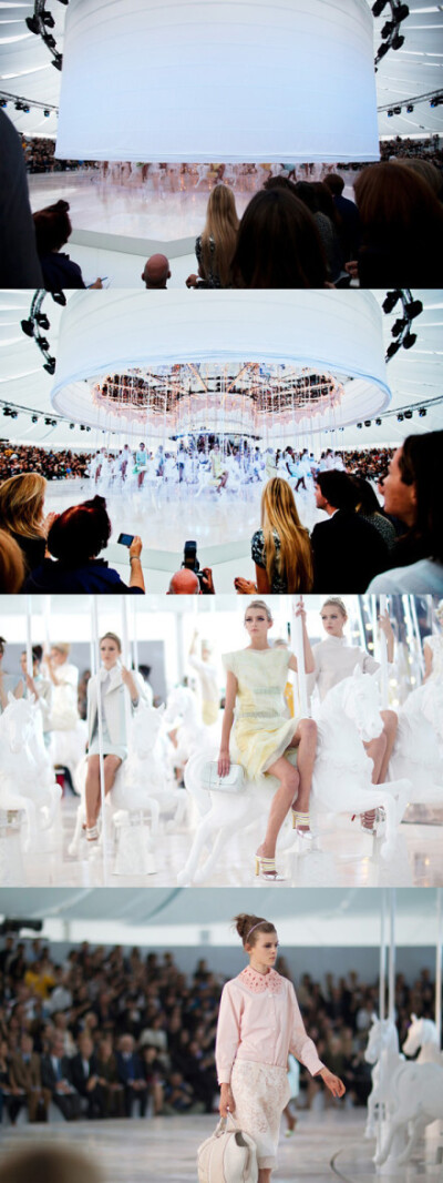 巴黎时装周Louis Vuitton 2012春季时装发布会开场，整体纯白色调，T台中心是一个白布包裹的巨型圆筒，布筒升起后露出一个众多模特坐着的巨大旋转木马。