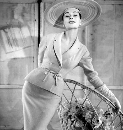 Anne Gunning is wearing double-breasted suit with tie at waist by Balmain, with broad brimmed hat, 1954