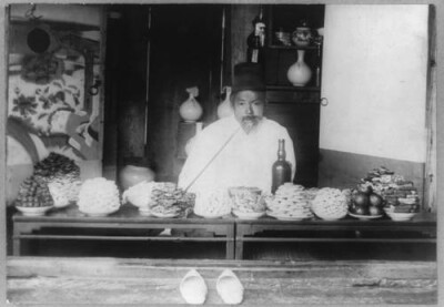 拍摄于1890年-1923年间，首尔一家食品店的店主。这位店主手持一柄极长的烟管站在食品店前，店里出售米糕、柿子和其他各类食品。尽管朝鲜人的服饰风格已今非昔比，但这些食物看上去熟悉依旧。