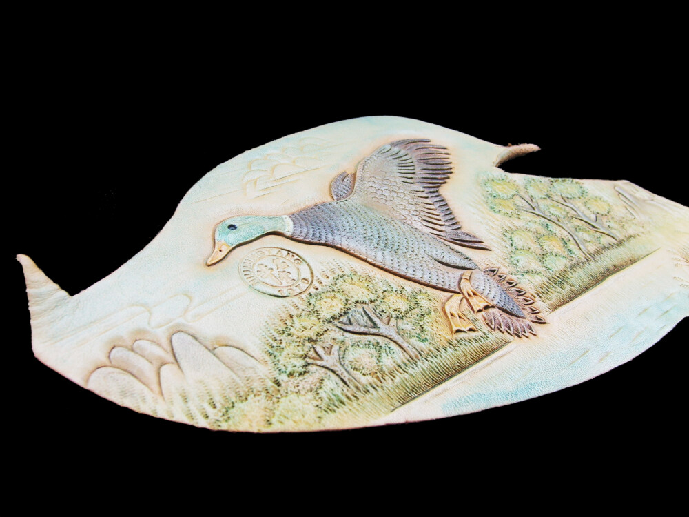 一只野鸭从水面扑棱跃起，手工雕刻染色，来自民间艺人的皮雕画手作。在展览上，他还会展出龙、虎皮雕画以及文具盒、鼠标垫等生活品皮雕