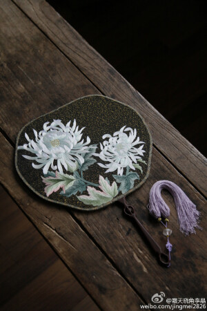 #团扇# 双面手绣团扇一款。黑色捻金线素缂丝底料，仿佛薄雾中浸出点点星光的夜空，细细勾勒出两朵雪菊，迎风而立，赛雪欺霜。花瓣间光影交叠，栩栩然跃于扇面，与清冷的老水晶珠子扇坠遥相呼应。红酸枝扇柄，真丝回龙须