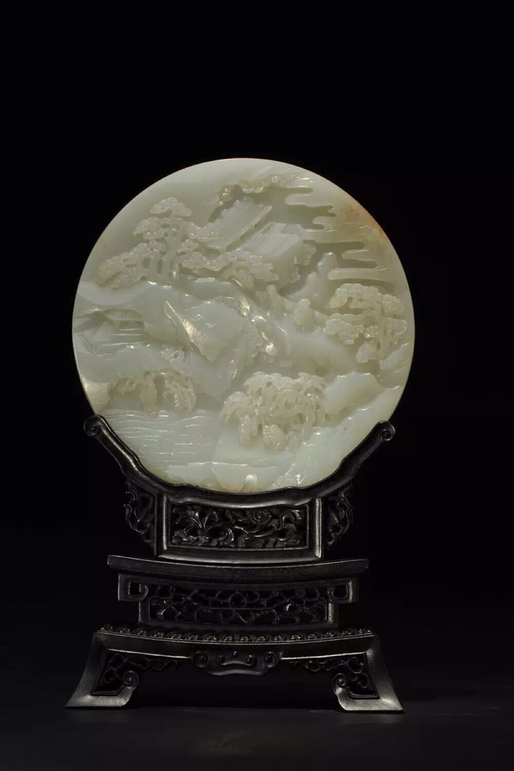 清乾隆 白玉壽山福海圓插屏帶紫檀座
直徑 D 14cm；高 H 22.8 cm
插屏為圓形片狀，玉質(zhì)凈白溫潤，色純則雅，下承鏤雕紫檀木座。一面頂部浮雕高山行云，云呈參差細(xì)長直條狀，頗具詩意。上部雕一長殿，中部橫亙一片巨大山石，嶙峋陡峭，上立松樹數(shù)棵，亭亭如蓋，兩位長者拾級(jí)而上于松蔭之下。下部山澗溪谷之中，云氣飄渺，松柏齊茂，隱約中有一亭臺(tái)掩映其間，一人坐于亭中舉目遠(yuǎn)眺。二人立于樹下汲水攀談，悠然自得。整體畫面不僅表現(xiàn)了山林洞府間的靜謐恬淡，更有仙山秘境飄渺不凡的氣質(zhì)。另一面浮雕汨汨江水邊一觀景樓臺(tái)，水流湍急，波浪翻涌，二童子雙手捧花立于欄邊，玩耍嬉鬧，言笑晏晏，身后古木參天，松虬盤纏，枝葉繁茂，雙童目光所至，空中五只蝙蝠振翅盤旋，寓意五福臨門。近方水邊山石之上，雕二仙鶴斂翼停立，使此屏面更加靈動(dòng)而富有生機(jī)，表達(dá)了仙福永享，壽與天齊的美好愿望。整器構(gòu)圖如畫，布局要妙，層次分明，剛?cè)嵯酀?jì)，虛實(shí)相合，靜水流深，遠(yuǎn)山如黛，于方寸之間展現(xiàn)出豐富景深，頗有意境，閑時(shí)賞玩，彷若置身其中，曲徑通幽處，漫步閑野間，足不出戶而可神游天地