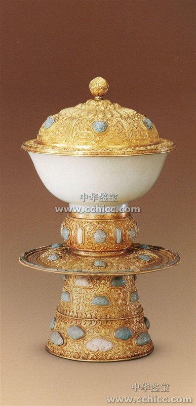 金錾花高足白玉盖碗 清,通高26厘米口径14.5厘米清宫旧藏 碗玉质，敞口，深腹，收底，圈足。碗外壁光素，内口沿镌刻藏文，碗配九成金盖，座。盖呈半圆形，顶配宝珠钮。盖面饰隐起莲瓣纹三层，卷草纹六组，每组内嵌松石雕花朵。座鼓形，锤錾隐起的卷草纹，嵌松石梅花云纹等。座下垫盘，盘面锤錾卷草纹，内嵌松石花朵，盘沿嵌长方形松石。盘下高撇足，外壁錾刻隐起的卷草纹云纹如意纹嵌梯形如意形梅花形松石嵌不同颜色、形状的松石46块。