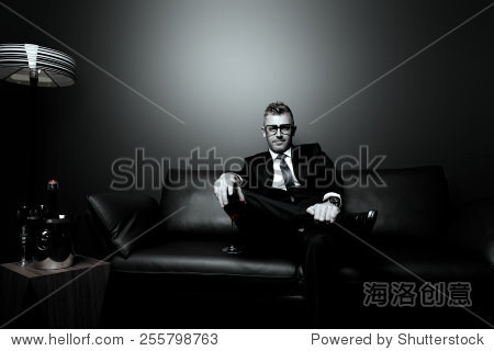 Black-and-white portrait of a handsome mature man in elegant suit drinking red wine. He is sitting on a leather couch in a luxurious interior.