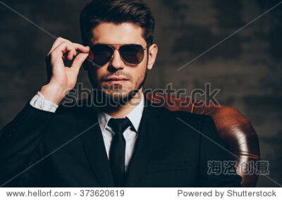 Portrait of confidence.Portrait of young handsome man in suit adjusting his sunglasses and looking at camera while sitting in leather chair against dark grey background