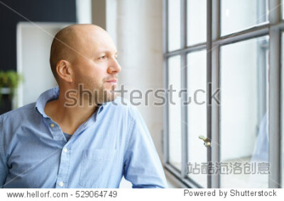 Middle-aged man watching through a window with a thoughtful expression as he leans on the sill