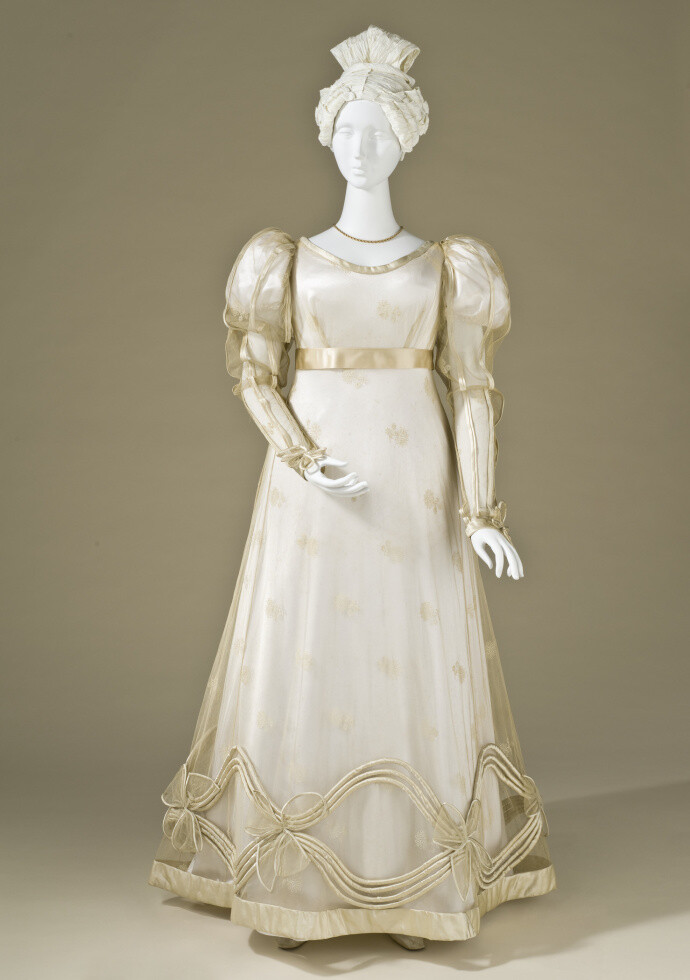 礼服，1825年，浪漫主义时代前期最流行的女装样式。一件白绸泡泡袖高腰长裙，外罩有细小花纹的长袖透明纱衣，各处均有象牙色丝绸镶边，裙摆上装饰着缎带和薄纱花结组成的花边。 ​​​​