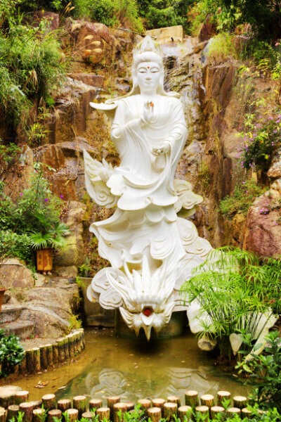 White Buddhist statue on background of small waterfall in a garden with pond