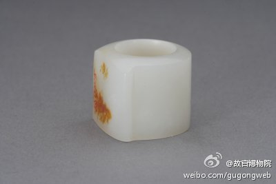 青白玉扳指，清，直径3.10cm，高2.80cm。清宫旧藏。扳指，又称“搬指”，是护指之物，一般多用玉做成指环状。最初戴于右手大拇指上，以免拉弓勾弦时勒手，后演化为一种装饰品。此扳指马鞍型，鞍面上有黄色桂花皮，玉质洁白莹润。附黄条，其上墨书“府脂玉皮搬指一个”。 ​​​​ …