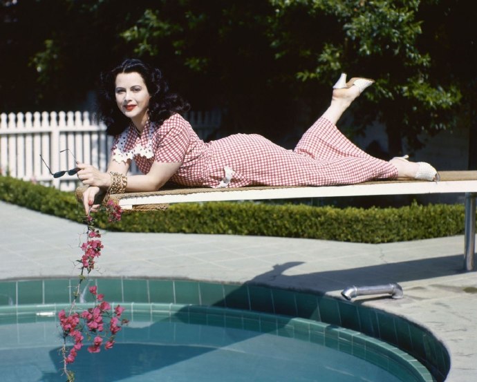 Hedy Lamarr wearing a red gingham dress and holding a branch with pink flowers while leaning, front down, on a diving board over the water of a swimming pool against velour-covered surface, 1945