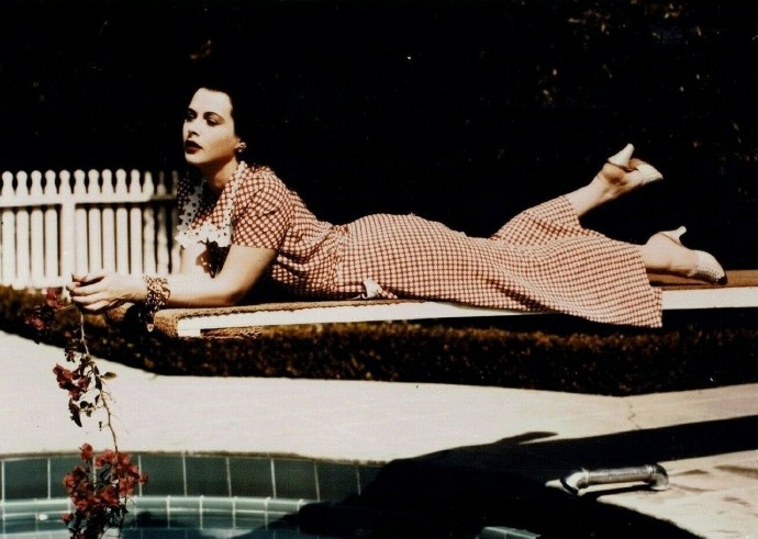 Hedy Lamarr wearing a red gingham dress and holding a branch with pink flowers while leaning, front down, on a diving board over the water of a swimming pool against velour-covered surface, 1945